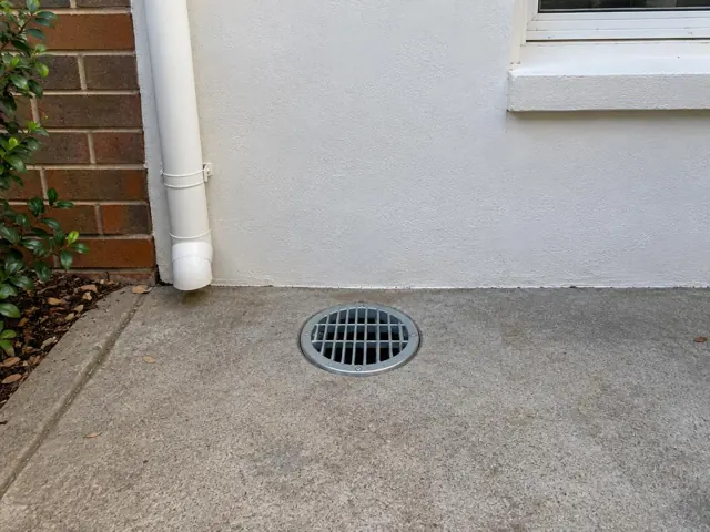 Outdoor yard drain grate set in dry concrete beside a white wall and downpipe in Roseville